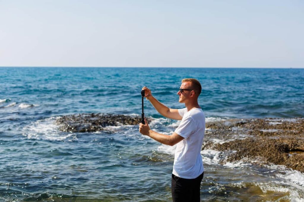 A man in a white T-shirt holds an action camera in his hands and shoots a beautiful view of the sea in barcelona in september