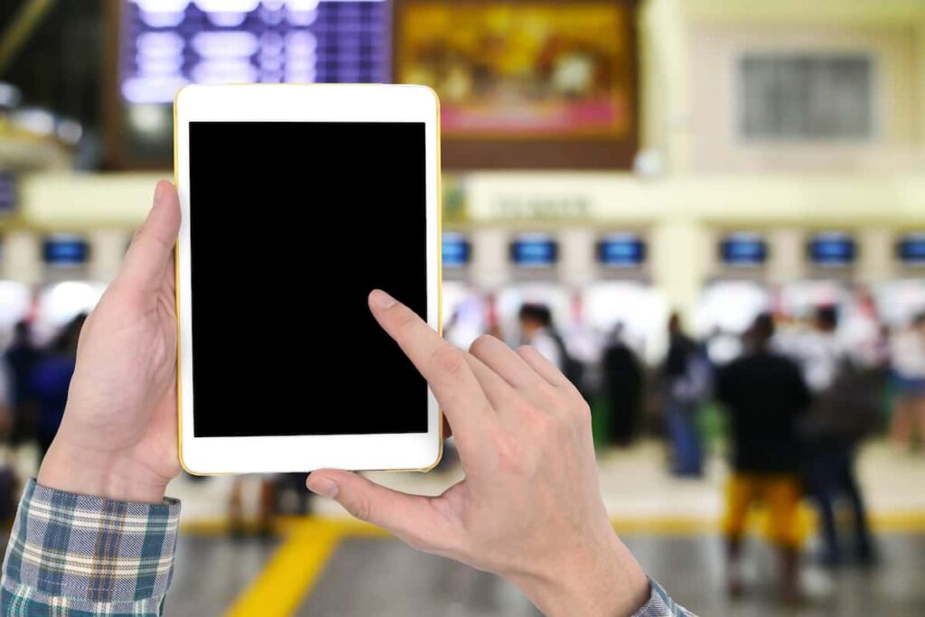 Male hands holding a white tablet with touch blank black screen on blurred image of people buy ticket at the counter background.