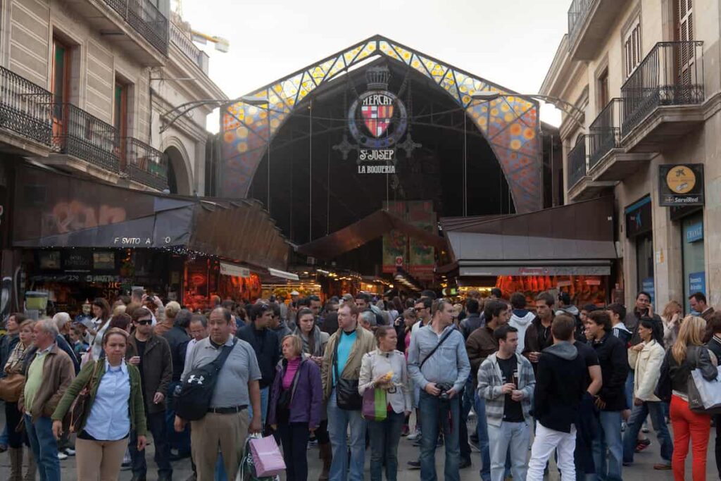 Crowd of famous La Boqueria market with Jamon, vegetables and fruits so that you can give Yourself a Few Days to See Everything is one of the best Barcelona travel tips.