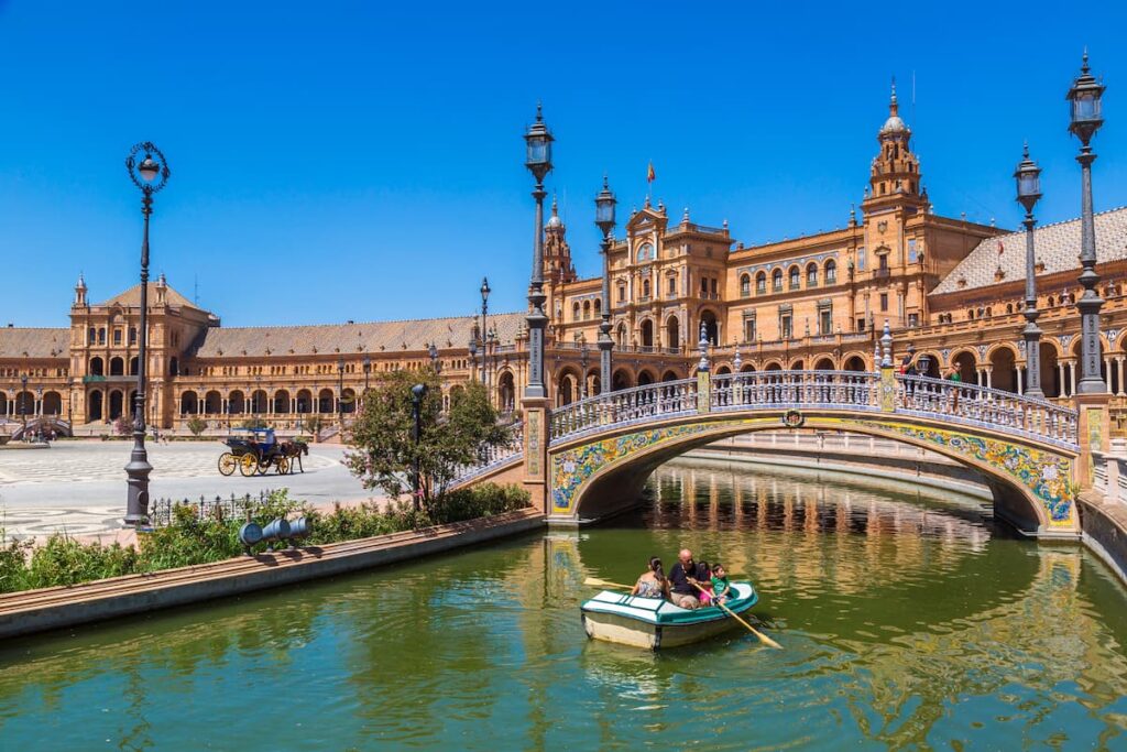A family rows a small boat along the canal in front of the ornate buildings and bridge at Plaza de España in Seville